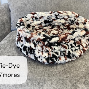 Puede incluir: Una cama redonda para mascotas te&ntilde;ida con tintes en tonos negros, blancos y marrones. La cama est&aacute; hecha de un material suave y afelpado y se encuentra sobre una superficie gris. El texto "Tie-Dye S'mores" est&aacute; en un rect&aacute;ngulo blanco.