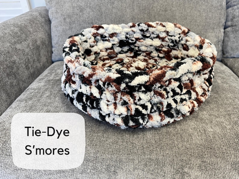 Puede incluir: Una cama redonda para mascotas te&ntilde;ida con tintes en tonos negros, blancos y marrones. La cama est&aacute; hecha de un material suave y afelpado y se encuentra sobre una superficie gris. El texto "Tie-Dye S'mores" est&aacute; en un rect&aacute;ngulo blanco.