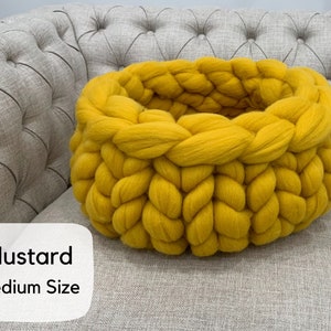 May include: A round, mustard yellow basket crafted from thick, chunky knit yarn. The basket features a prominent braided texture throughout its circular structure. A white label on the lower left displays the text Mustard Medium Size.