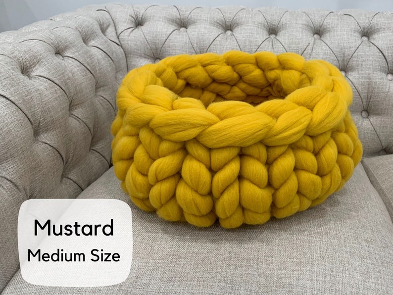 May include: A round, mustard yellow basket crafted from thick, chunky knit yarn. The basket features a prominent braided texture throughout its circular structure. A white label on the lower left displays the text Mustard Medium Size.