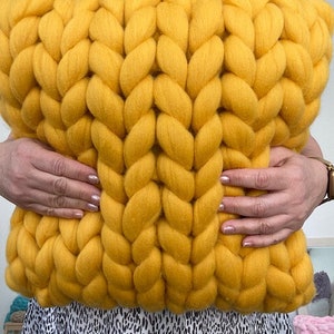 May include: A large, yellow knitted throw pillow with a chunky cable knit pattern. The pillow is being held by two hands.
