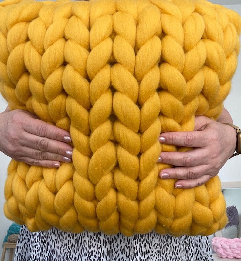 May include: A large, yellow knitted throw pillow with a chunky cable knit pattern. The pillow is being held by two hands.