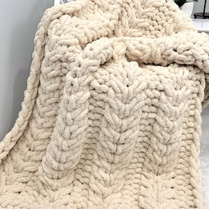 May include: A chunky knit blanket in a solid cream color, featuring a thick, soft yarn texture with a prominent braided stitch pattern. The blanket is draped over a white chair, showcasing its heavy, voluminous, and cozy appearance.