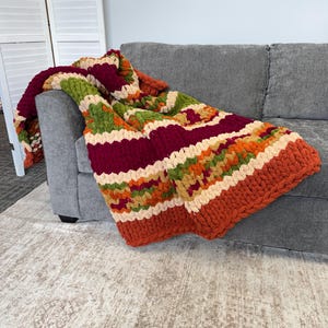 Op de afbeelding: Een grof gebreide plaid met horizontale strepen in afwisselende kleuren van diep bordeauxrood, olijfgroen, oranje en cr&egrave;me. De deken is gemaakt van dik, getextureerd garen in een herhalend patroon dat over de armleuning van een bank valt.