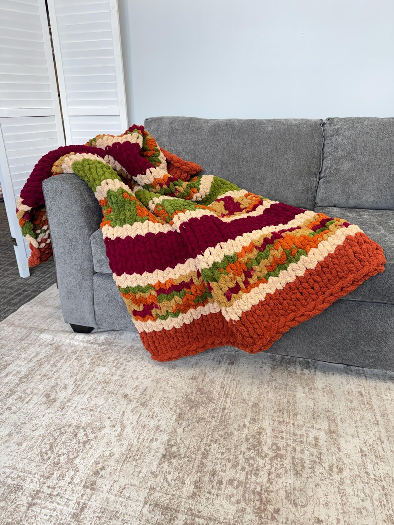 Op de afbeelding: Een grof gebreide plaid met horizontale strepen in afwisselende kleuren van diep bordeauxrood, olijfgroen, oranje en cr&egrave;me. De deken is gemaakt van dik, getextureerd garen in een herhalend patroon dat over de armleuning van een bank valt.