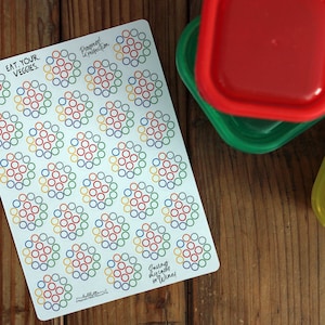 Può includere: Un foglio di adesivi con la scritta "EAT YOUR VEGGIES" e disegni circolari colorati. Accanto agli adesivi ci sono quattro contenitori per alimenti in plastica con coperchi rossi, verdi, blu e arancioni.