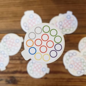 May include: A collection of white, circular stickers with a colorful design. Each sticker features a central pattern of colored circles, including red, blue, green, and yellow. The stickers are arranged on a wooden surface.