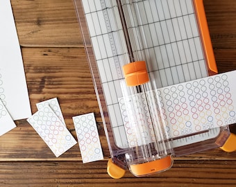 May include: A close-up of a paper cutter with a roll of colorful circle stickers being cut. The stickers have a white background with red, yellow, blue, and green circles.