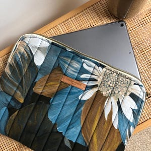 Peut inclure: Une housse d'ordinateur portable matelassée avec un motif floral dans les tons bleus, bruns et blancs. La housse est dotée d'une fermeture éclair et d'une étiquette en cuir marron avec le texte "MaisonLabo Paris".
