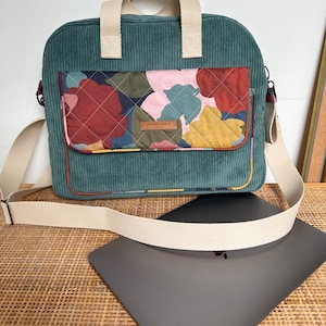 Peut inclure: Sac d'ordinateur portable en velours côtelé vert d'eau avec une poche florale matelassée rouge, rose et jaune. Le sac a une poignée et une bandoulière crème. Un ordinateur portable gris repose sur une surface tissée.