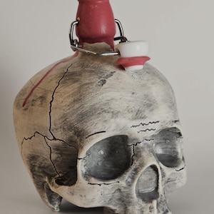 May include: A skull-shaped bottle with a weathered off-white and grey finish featuring dark cracks and a red streak. The bottle has a pink neck and a metal swing-top closure with a white stopper. The design includes black line details.
