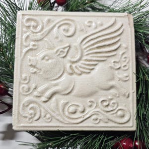 May include: Off-white square ceramic tile featuring a relief of a winged pig in flight. The tile has a crackled surface and decorative scrollwork around the edges. A whimsical, decorative item.
