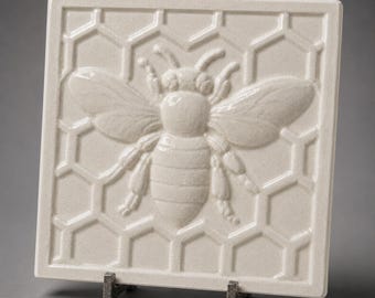 May include: A square, off-white ceramic tile featuring a raised, embossed design of a honeybee centered over a geometric hexagonal honeycomb pattern. The tile has a glossy finish and is supported by a small, dark metal stand at the base.