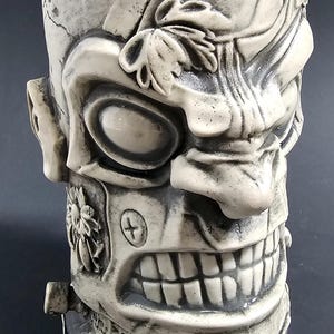 May include: A detailed, off-white ceramic tiki mug with a monster face design. The mug features a large, toothy grin, a prominent nose, and stylized facial features. Decorative elements include a flower and a bolt.