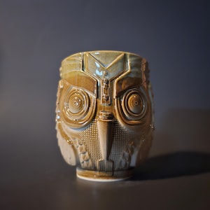 Może przedstawiać: Ceramiczny kubek w kształcie sowy z brązową i niebieską glazurą. Sowa ma szczegółowe oczy i dziób.