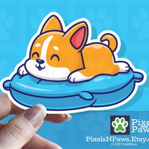 May include: A sticker featuring a cartoon corgi dog lying on a blue pillow. The corgi is orange and white with a happy expression. The background is light blue with paw prints. The sticker is held by a hand.