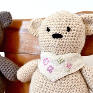 May include: Two crocheted teddy bears, one brown and one beige, are sitting on a wooden chair. Both bears are wearing a white fabric bib with pink and green embroidered flowers.