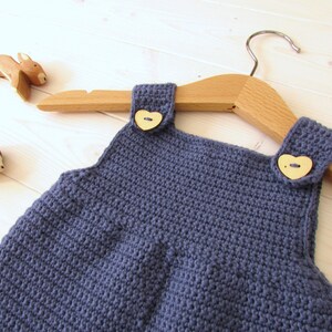 May include: A blue crocheted baby overall with wooden heart-shaped buttons. The overall is hanging on a wooden hanger.