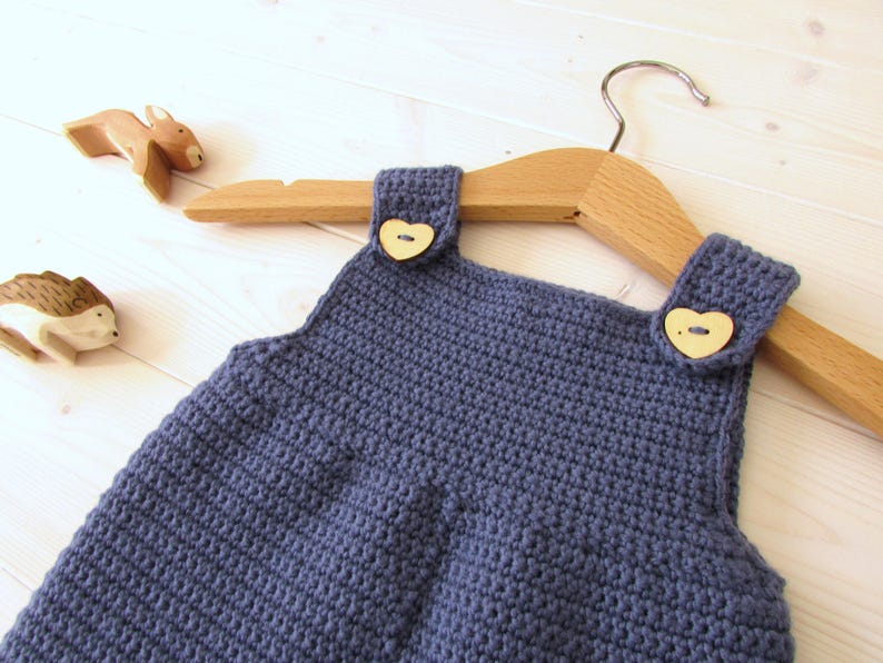 May include: A blue crocheted baby overall with wooden heart-shaped buttons. The overall is hanging on a wooden hanger.
