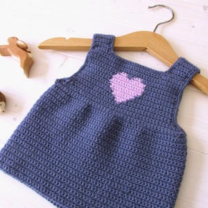 May include: A blue crocheted sleeveless dress with a pink heart on the front. The dress is hanging on a wooden hanger.