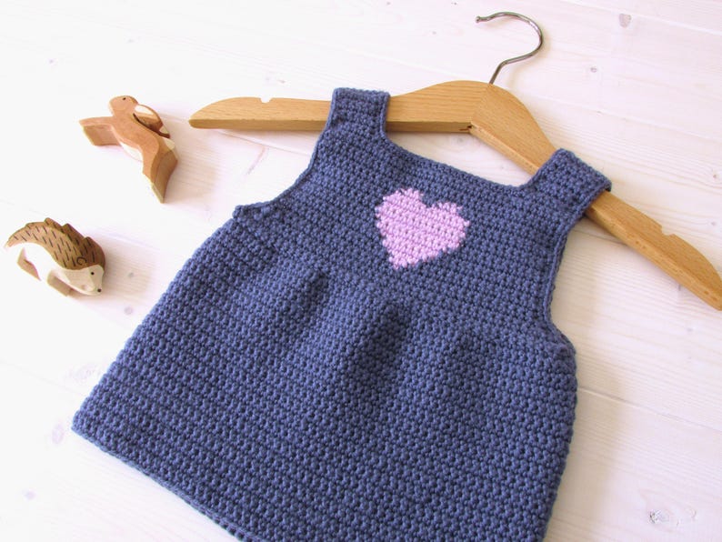May include: A blue crocheted sleeveless dress with a pink heart on the front. The dress is hanging on a wooden hanger.