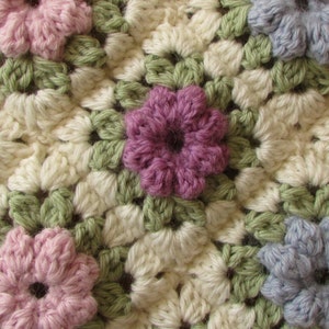 May include: A close-up of a crocheted blanket featuring a repeating pattern of flowers in shades of pink, purple, and blue. The flowers are surrounded by white and green crocheted squares.