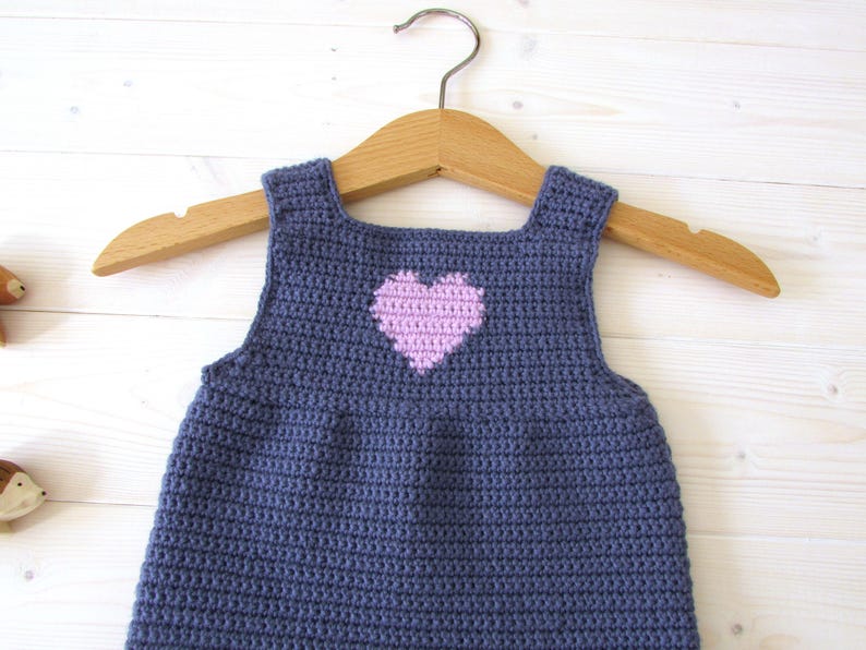 May include: A crocheted baby dress in a dark blue color with a pink heart design on the front. The dress is hanging on a wooden hanger.