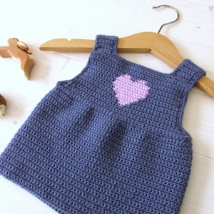 May include: A blue crocheted sleeveless dress with a pink heart design. The dress is hanging on a wooden hanger.