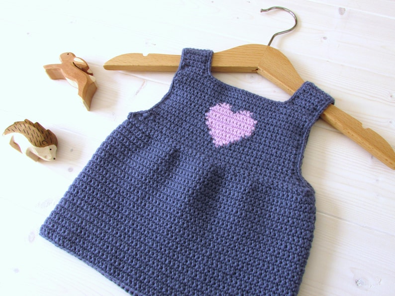 May include: A blue crocheted sleeveless dress with a pink heart design. The dress is hanging on a wooden hanger.