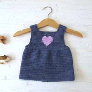 May include: A sleeveless crocheted baby dress in a dark blue color with a pink heart on the front. The dress is hanging on a wooden hanger.