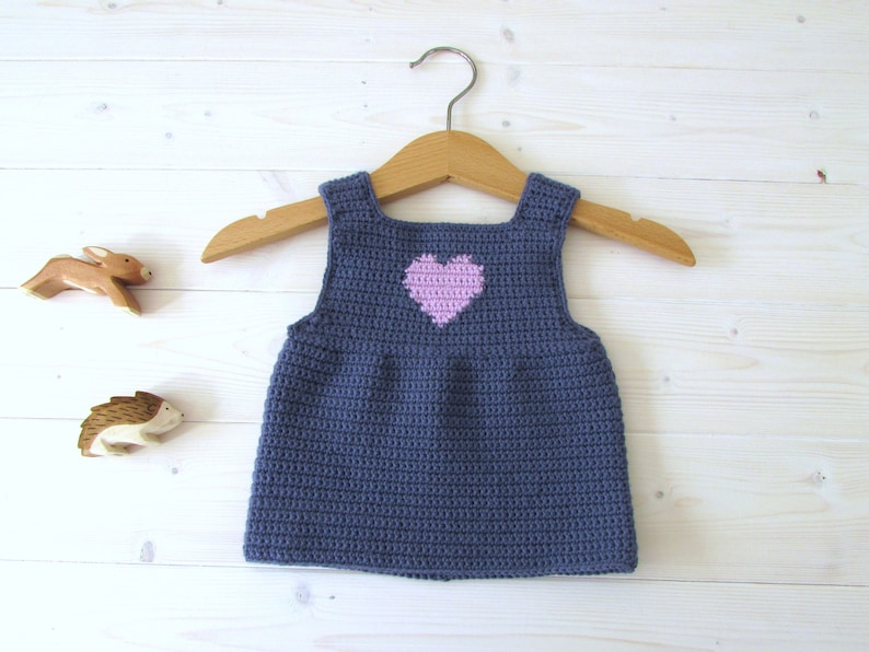 May include: A sleeveless crocheted baby dress in a dark blue color with a pink heart on the front. The dress is hanging on a wooden hanger.