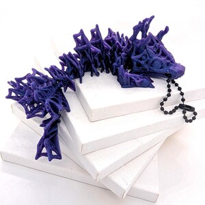 May include: A detailed, articulated purple dragon figurine with a textured surface. The dragon is lying on a stack of white canvases. A black beaded chain is attached to the dragon's head.