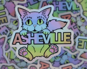 May include: A holographic sticker featuring a cartoon cat holding a sign that says "Asheville". The cat is green, blue, and purple with a pink nose and paws. The sign is rainbow colored.