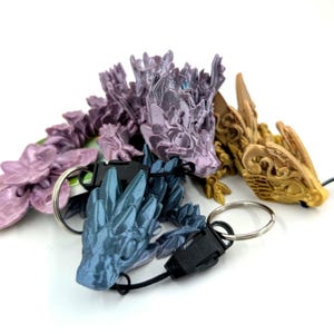 May include: A collection of 3D-printed dragon keychains in shades of purple, blue, and gold. Each dragon has intricate details and is attached to a black clasp and a silver keyring. The keychains are arranged on a white surface.