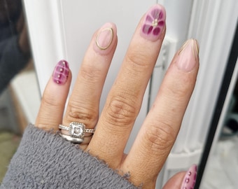 Puede incluir: Una mano con esmalte de uñas rosa y dorado. Las uñas están decoradas con pequeñas flores y puntos. La mano lleva un anillo de plata con un diamante de forma cuadrada.