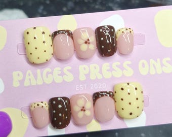 Puede incluir: Un conjunto de uñas postizas con varios diseños. Algunas uñas son amarillas con lunares marrones, otras rosas con una flor blanca y otras marrones con lunares blancos. El respaldo rosa muestra el texto "PAIGES PRESS ONS EST 2020".