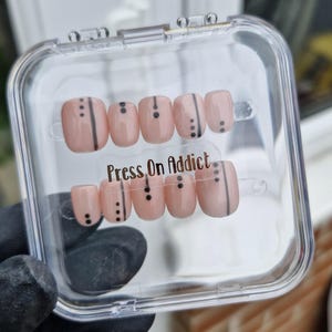 May include: A clear plastic case containing a set of 10 press-on nails. The nails are a light pink colour with black lines and dots. The case has the text "Press On Addict" printed on it.
