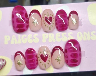 Puede incluir: Un conjunto de uñas postizas con varios diseños. Algunas uñas son de color magenta liso, mientras que otras presentan un patrón magenta. Varias uñas tienen una base nude con puntas magenta y detalles dorados, incluyendo corazones y estrellas. El texto "PAIGES PRESS ONS" es visible.