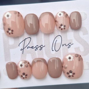 May include: A collection of oval press-on nails in shades of pink and brown. Some nails are decorated with a small white flower design with brown centres. The words "Press Ons" are written in cursive.