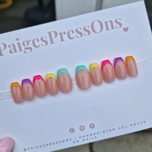 May include: A set of ten press-on nails with a rainbow coloured French tip design. The nails are on a white card with the text "PaigesPressOns."