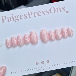 May include: Pink oval-shaped press-on nails with white polka dots. The nails are arranged in a row on a white background. The text "PaigesPressOns." is at the top of the image. The text "@paigespressons | handpainted gel press on nails" is at the bottom of the image.