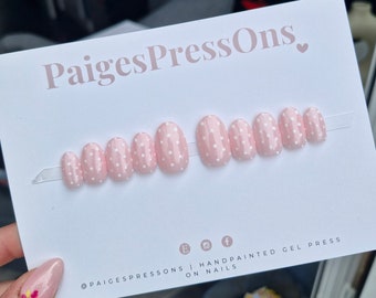 Puede incluir: Uñas postizas ovaladas de color rosa con lunares blancos. Las uñas están dispuestas en una fila sobre un fondo blanco. El texto "PaigesPressOns." está en la parte superior de la imagen. El texto "@paigespressons | handpainted gel press on nails" está en la parte inferior de la imagen.
