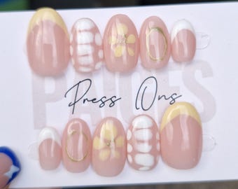 Puede incluir: Un conjunto de diez uñas postizas con un diseño floral amarillo y blanco. Las uñas son de forma ovalada y tienen una base rosa claro. El texto "Press Ons" está escrito en negro sobre un fondo blanco.