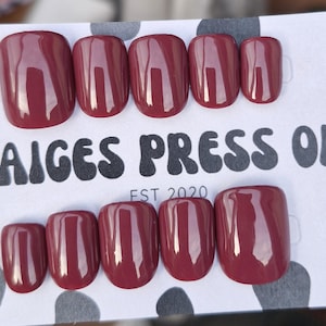 May include: A set of ten glossy, dark mauve press-on nails arranged in two rows on a white display card with a black cow print pattern. The card features the text PAIGES PRESS ONS and EST 2020 in a bold, rounded black font centred between the nail rows.