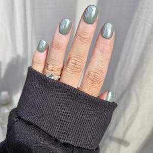 May include: A hand with short, oval-shaped nails painted a shimmery green colour.