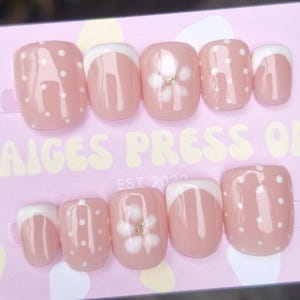 May include: A set of pink press-on nails with white polka dots and a white French tip design. Some nails feature a white flower design with a gold centre. The text "PAIGES PRESS ONS EST 2020" is visible on the pink background.