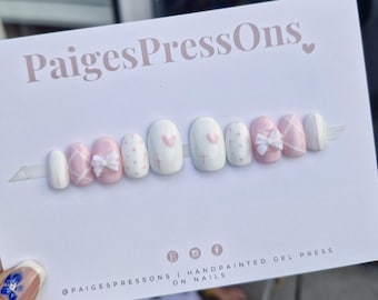 May include: Set of ten white and pink press-on nails with various designs, including polka dots, hearts, bows, and a plaid pattern. The nails are displayed on a white card with the text "PaigesPressOns."