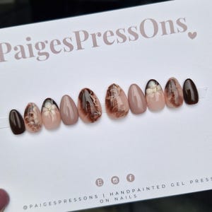 May include: A collection of oval-shaped, hand-painted gel press-on nails. The designs include solid dark brown, floral patterns, and marble effects in brown, beige, and white. The card displays the text "PaigesPressOns".