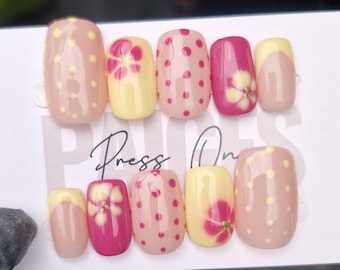 Puede incluir: Un conjunto de uñas postizas con varios diseños. Algunas uñas son de color rosa claro con lunares amarillos, otras son amarillas con diseños florales rosas, y algunas son de color rosa intenso. El texto "Press On" es visible en la tarjeta.