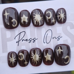 May include: Dark brown press-on nails with gold celestial designs, including stars, moons, and suns. The nails are arranged on a white card with the words "Press Ons" in black script.